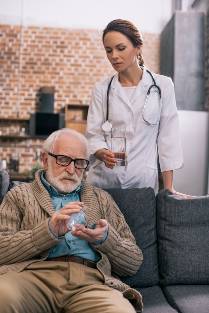 Starszy mężczyzna siedzący na sofie i przyjmujący lekarstwo. Stojąca przy nim opiekunka ze szklanką wody.