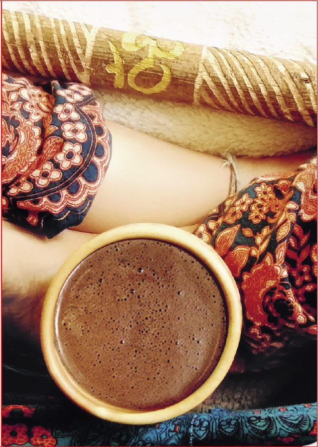 Kubek gęstego kakao ceremonialnego trzymany na skrzyżowanych nogach w kolorowych, etnicznych spodniach, obok drewnianej, rzeźbionej pałki z ornamentem. Zdjęcie pochodzi z książki "Kakao ceremonialne - napój otwierający serce. Przepisy, rytuały i praktyki duchowe, które wprowadzają spokój i pogłębiają świadomość" autorstwa
Felix Eidner Leni Glapa