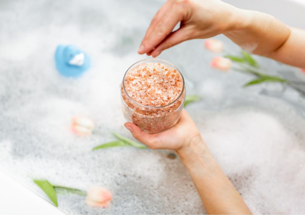 Kąpiel lecznicza w domu. Dłoń wsypuje kryształy soli mineralnych do wanny napełnionej wodą pianą i kwiatami.